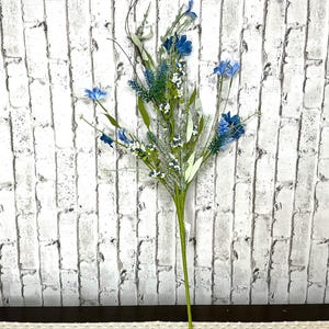 May include: A tall, artificial floral arrangement featuring blue flowers, green leaves, and small white accents. The stems are green, and the arrangement is set against a white brick wall background. This decorative piece is suitable for home decor.