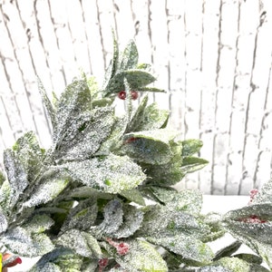 Puede incluir: Primer plano de una planta artificial verde y blanca con peque&ntilde;as bayas rojas. Las hojas est&aacute;n cubiertas de un polvo blanco, creando una apariencia helada. El fondo es una pared de ladrillos blancos.