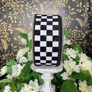 May include: A roll of black and white checkered ribbon. The ribbon is displayed on a white pedestal, surrounded by white flowers and green leaves. The background features a black and gold floral pattern.