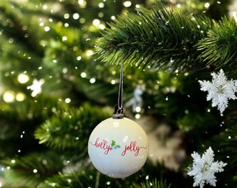 Puede incluir: Una bola de Navidad blanca con las palabras "holly jolly" en escritura roja y acentos de hojas de acebo. La bola cuelga de un hilo negro sobre un fondo de un árbol de Navidad verde con decoraciones de copos de nieve.