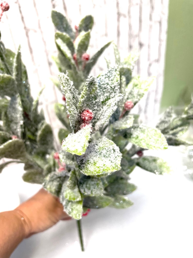 Puede incluir: Un ramo de vegetaci&oacute;n artificial con hojas verdes y peque&ntilde;as bayas rojas. Las hojas est&aacute;n espolvoreadas con una sustancia blanca y brillante, d&aacute;ndoles una apariencia helada. Los tallos son verdes.