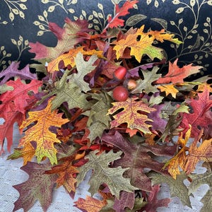 May include: A vibrant arrangement of artificial autumn leaves in shades of red, orange, yellow, and green. The leaves are oak-shaped and clustered together, with small red berries interspersed throughout. The background is a dark surface with gold floral accents.