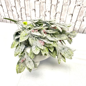 Puede incluir: Un ramo de vegetaci&oacute;n artificial con hojas verdes y peque&ntilde;as bayas rojas, espolvoreado con una sustancia blanca similar a la nieve. El arreglo se exhibe sobre un soporte para pasteles blanco, contra una pared de ladrillos blancos.