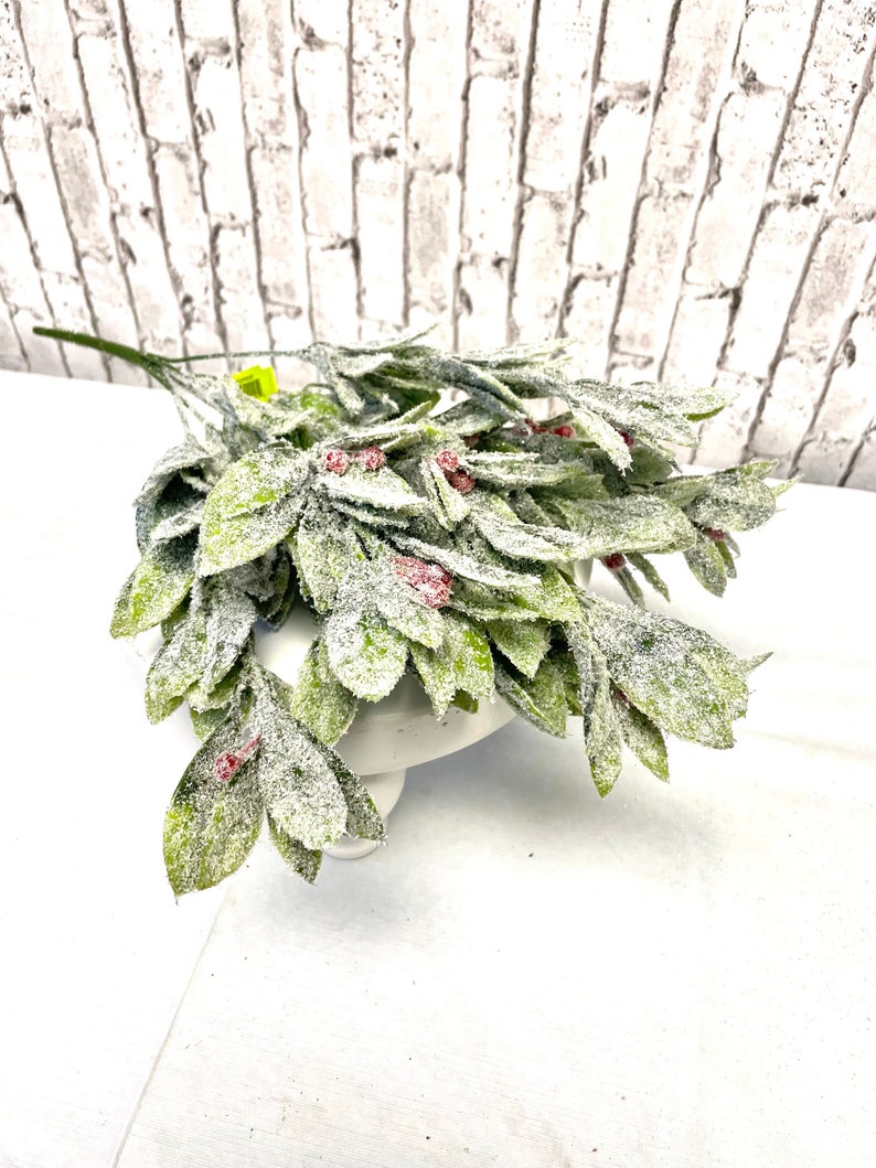 Puede incluir: Un ramo de vegetaci&oacute;n artificial con hojas verdes y peque&ntilde;as bayas rojas, espolvoreado con una sustancia blanca similar a la nieve. El arreglo se exhibe sobre un soporte para pasteles blanco, contra una pared de ladrillos blancos.