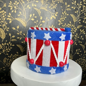 May include: A small, decorative drum with a patriotic theme. The drum features a blue top and bottom with white stars, red and white stripes, and red accents. White rope detailing and red beads add to the festive design.