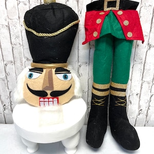 May include: A whimsical Christmas decoration featuring a nutcracker head atop a white stool and a pair of elf legs. The nutcracker has a black hat, gold accents, and a smiling face. The elf legs are green with a red jacket and black boots.