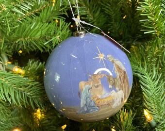 May include: A spherical Christmas ornament with a light blue background featuring a painted nativity scene. The illustration depicts figures, a cow, and a donkey gathered around a manger under a bright star. It hangs from a silver cap.