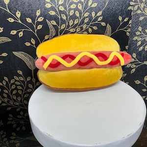 May include: A plush hot dog with a yellow bun, a pink sausage, and yellow and red mustard. The hot dog is sitting on a white pedestal against a black background with a gold floral pattern. A fun food-themed item.