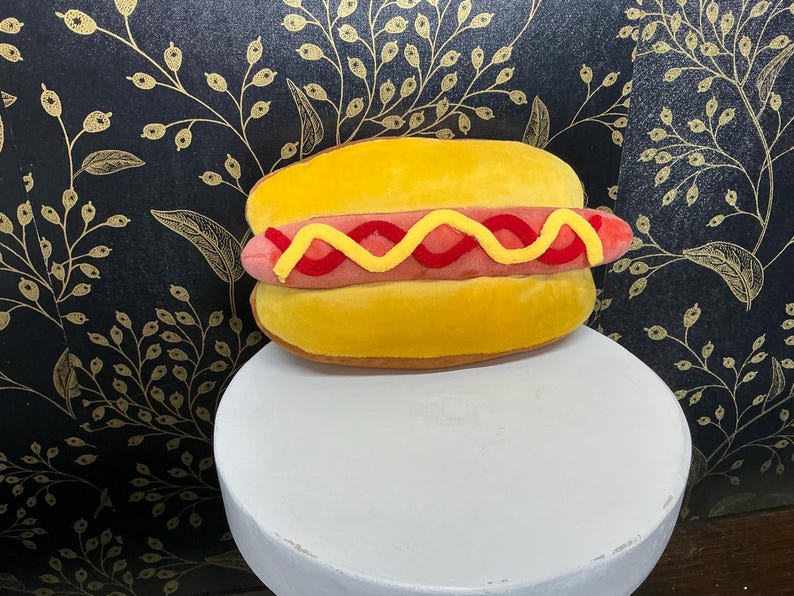 May include: A plush hot dog with a yellow bun, a pink sausage, and yellow and red mustard. The hot dog is on a white pedestal against a black background with a gold floral pattern. A fun food-themed item.