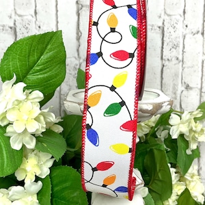 May include: White ribbon with a repeating pattern of colorful Christmas lights. The ribbon has a red border and is displayed against a backdrop of white flowers and green leaves. The lights are in red, yellow, green, blue, and orange.