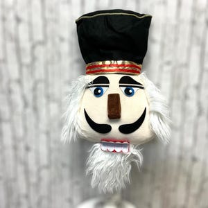 May include: A plush nutcracker head with a black hat, gold trim, and a red band. The face is cream-colored with blue eyes, a brown nose, black mustache, and a white beard. The mouth has a red and white tooth design.