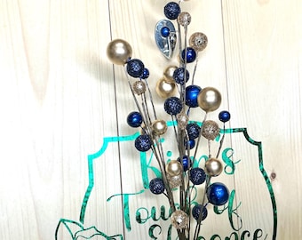 May include: Decorative floral arrangement featuring gold, navy blue, and glittery spheres on slender stems. The arrangement is set against a light wood background with a teal and green floral design that reads "Touch of Elegance."