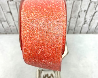 Puede incluir: Un carrete de cinta de color coral con una textura brillante. La cinta se muestra sobre un soporte plateado sobre un fondo de pared de ladrillo blanco. La cinta probablemente se utiliza para manualidades, envolver regalos o decoraciones.