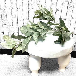 May include: A white, three-legged decorative stand with a round top, holding a sprig of green and gray faux lamb's ear leaves. The leaves have a soft, velvety texture. The stand is placed on a white, textured surface.
