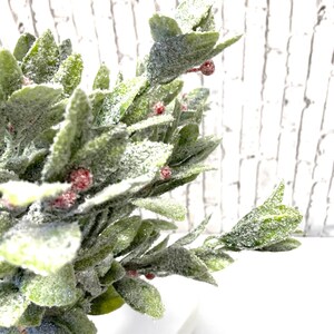 Puede incluir: Primer plano de una planta artificial decorativa con hojas verdes y peque&ntilde;as bayas rojas, espolvoreadas con una capa de escarcha blanca. La planta est&aacute; en una maceta blanca de estilo pedestal, creando una est&eacute;tica festiva y invernal.