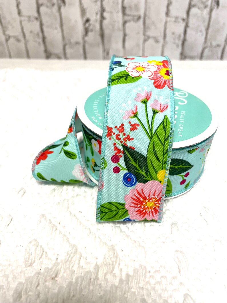 May include: A roll of light blue floral ribbon with a colorful flower pattern. The ribbon features pink, red, yellow, and white flowers with green leaves. The ribbon is on a white surface.