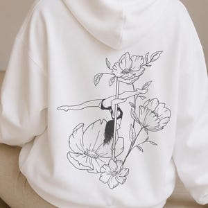 May include: A white hooded sweatshirt featuring a black line art graphic on the back. The design depicts a person performing a pole dance move surrounded by large floral illustrations. The hoodie has long sleeves and a hood, with a clean white finish.