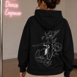 May include: A black hooded sweatshirt featuring a white line art graphic on the back. The design depicts a person performing a pole dance move integrated with large floral illustrations. The hoodie has long sleeves and a hood with a drawstring.