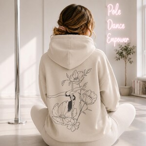May include: A beige hooded sweatshirt featuring a black line art graphic on the back. The design depicts a person performing a pole dance move integrated with large floral illustrations. The hoodie has long sleeves and a hood, worn over matching pants.