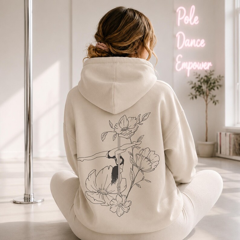 May include: A beige hooded sweatshirt featuring a black line art graphic on the back. The design depicts a person performing a pole dance move integrated with large floral illustrations. The hoodie has long sleeves and a hood, worn over matching pants.