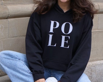 May include: Black hooded sweatshirt with the word "POLE" printed in white capital letters. The sweatshirt is worn with light blue jeans and white sneakers. The person is sitting on a concrete surface.
