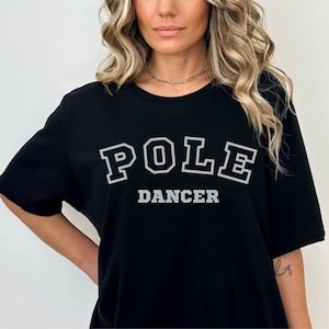 Puede incluir: Camiseta negra con el texto "POLE DANCER" en letras blancas con contorno.