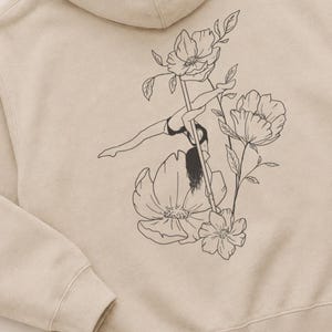 May include: A beige hooded sweatshirt featuring a black line art graphic on the back. The illustration depicts a person performing a pole dance move on a vertical pole, surrounded by large, detailed floral blossoms and leaves.