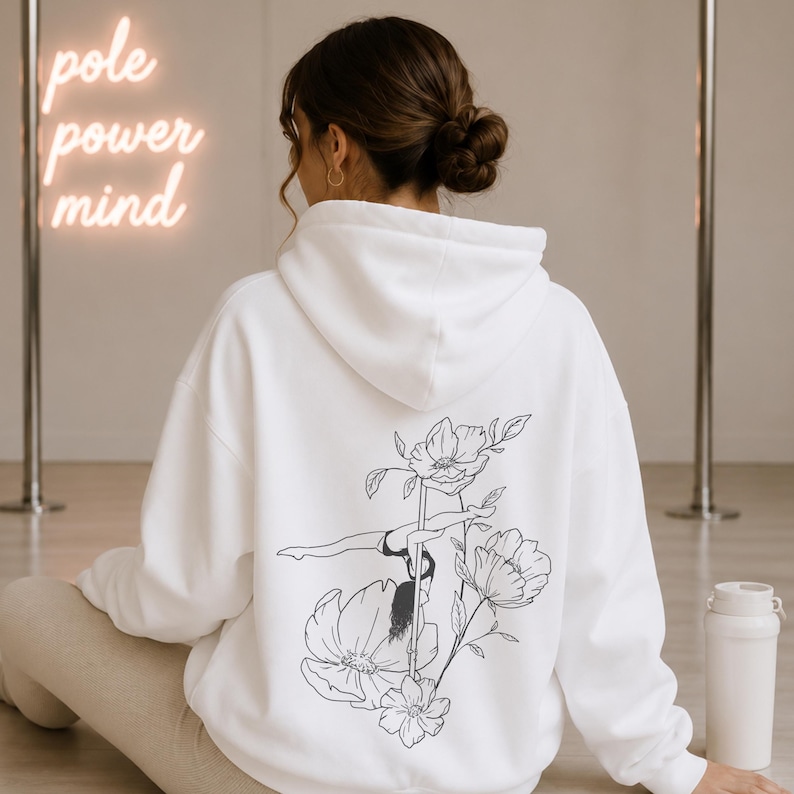 May include: A white hooded sweatshirt featuring a black line art graphic on the back. The design depicts a person performing a pole dance move surrounded by large floral illustrations. The hoodie has long sleeves and a hood, with a clean white finish.