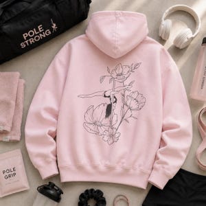 May include: A light pink hooded sweatshirt featuring a black line art graphic on the back. The design depicts a person performing a pole dance move integrated with floral illustrations. The hoodie has long sleeves, a hood, and ribbed cuffs.