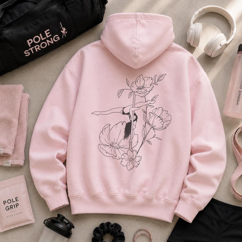 May include: A light pink hooded sweatshirt featuring a black line art graphic on the back. The design depicts a person performing a pole dance move integrated with floral illustrations. The hoodie has long sleeves, a hood, and ribbed cuffs.