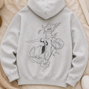 May include: A light gray hooded sweatshirt featuring a black line art graphic on the back. The design depicts a person performing a pole dance move, surrounded by large, detailed floral illustrations of blooming flowers and leaves.