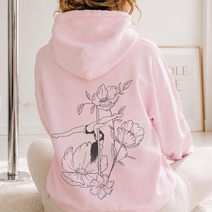 May include: A light pink hooded sweatshirt featuring a black line art graphic on the back. The design depicts a person performing a pole dance move integrated with large floral illustrations. The hoodie has long sleeves and a hood.