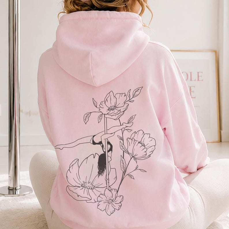 May include: A light pink hooded sweatshirt featuring a black line art graphic on the back. The design depicts a person performing a pole dance move integrated with large floral illustrations. The hoodie has long sleeves and a hood.