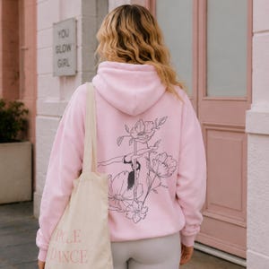 May include: A light pink hooded sweatshirt featuring a black line art graphic on the back. The design depicts a person performing a pole dance move surrounded by large floral illustrations. A beige tote bag with the text POLE DANCE is carried.