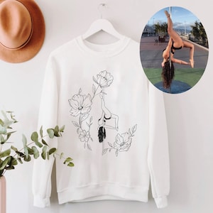 May include: White crew neck sweatshirt with a black line drawing of a pole dancer surrounded by flowers. An inset image shows a person performing the same pole dance. A brown hat and green plant are in the background.