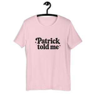 Op de afbeelding: Een lichtroze T-shirt met korte mouwen en de zwarte tekst "Patrick told me" op het midden van de borst. De tekst is geschreven in een vet, afgerond lettertype en wordt geaccentueerd door vier kleine, vierpuntige sterpictogrammen rondom.