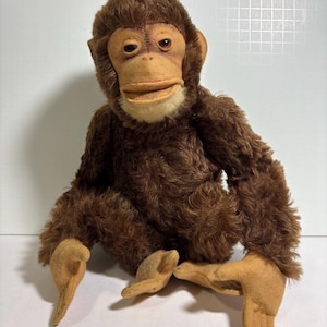 Könnte beinhalten: Ein Vintage-Stoffaffe mit braunem Fell und einem beigefarbenen Gesicht, Händen und Füßen. Der Affe hat einen detaillierten Gesichtsausdruck mit dunklen Augen und einem lächelnden Mund. Das Plüschtier sitzt aufrecht, mit ausgestreckten Armen und Beinen.