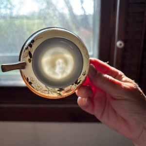 May include: An antique teacup, held up to the light, revealing its interior. The cup features a gold handle and rim, with floral and foliage designs in pink, yellow, and black. The cup's interior is a light gray color.