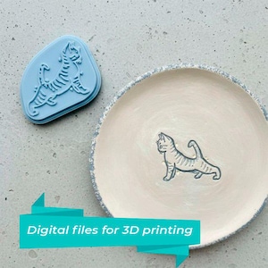 May include: A light blue cat-shaped stamp and a ceramic plate with a matching cat design. The plate has a cream-colored surface with a blue rim. The text "Digital files for 3D printing" is displayed on a teal banner.