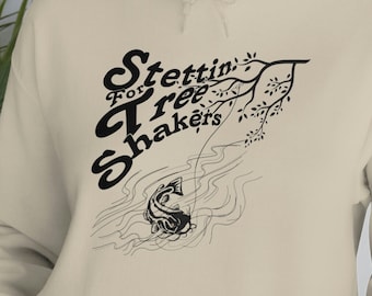 May include: A beige hoodie with a black graphic print of a fishing scene. The text on the graphic reads "Stettin for Tree Shakers".