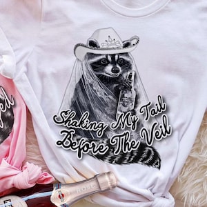 May include: Two t-shirts, one pink and one white, with raccoon graphics. The pink shirt says "Feral Friends Unhinged." The white shirt says "Shaking My Tail Before The Veil." A bottle of rose wine is in the foreground. Floral arrangement in the background.