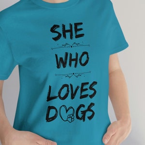 Peut inclure: Un t-shirt bleu turquoise avec le texte "SHE WHO LOVES DOGS" imprim&eacute; en noir. Le texte est stylis&eacute; avec un c&oelig;ur et une empreinte de patte dans le mot "DOGS".