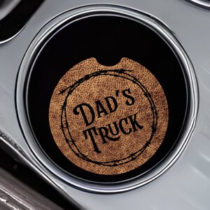 May include: Two brown leather car coasters. One coaster features a black and white illustration of a cow and calf. The other coaster has a brown circle with the text "Dad's Truck" in black lettering.