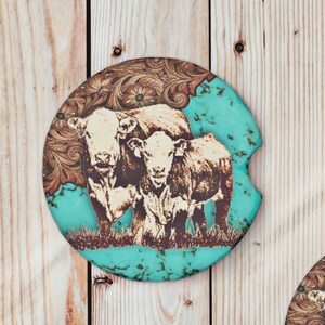 May include: Two turquoise blue coasters with a brown floral design and white cows. The cows are standing in a field of grass.