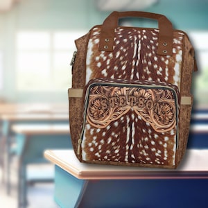 May include: A brown leather backpack with a deer print design and a tooled leather pocket with the word "Stetson" embossed on it. The backpack is sitting on a wooden desk.