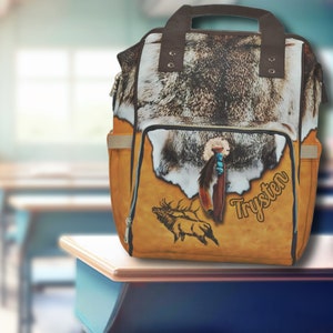 May include: A brown and tan backpack with a faux fur flap and a brown leather handle. The backpack has a large pocket on the front with a brown elk design, a turquoise and brown feather design, and the name "Trysten" written in brown.