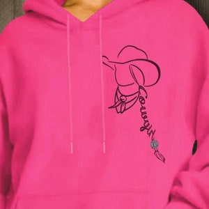 May include: Bright pink hoodie with a kangaroo pocket and drawstring hood. A black line drawing of a cowboy hat and the word "Cowgirl" is printed on the front. The design includes a turquoise-colored accent.