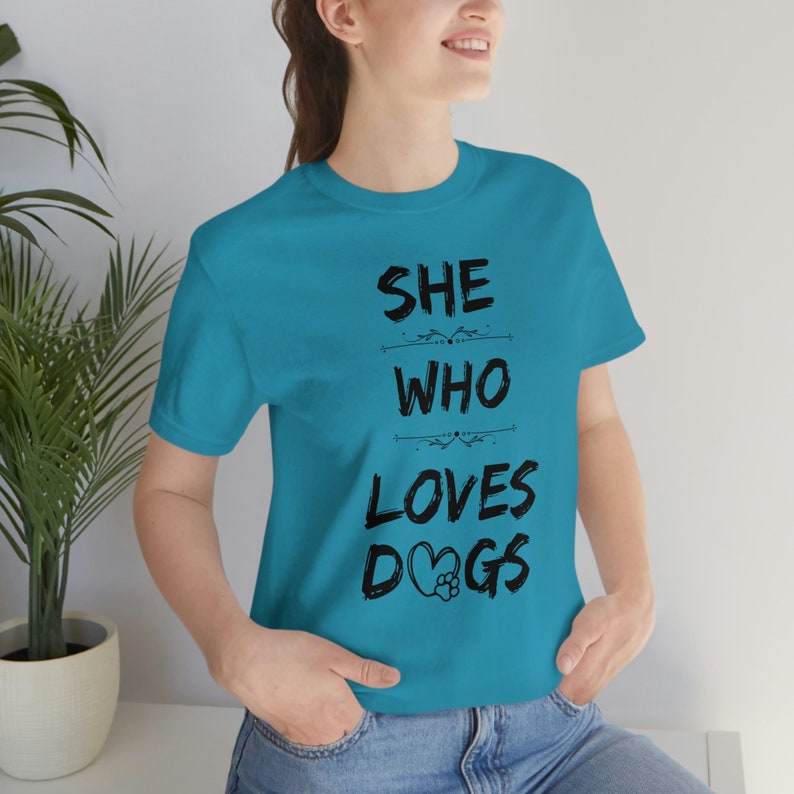 Peut inclure: Un t-shirt bleu turquoise avec le texte "SHE WHO LOVES DOGS" imprim&eacute; en noir. Le texte est stylis&eacute; avec un c&oelig;ur et une empreinte de patte dans le mot "DOGS".