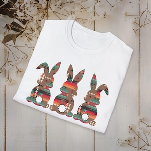 May include: White t-shirt with three brown bunny silhouettes. The bunnies are patterned with colorful geometric designs in shades of red, orange, blue, green, and white.