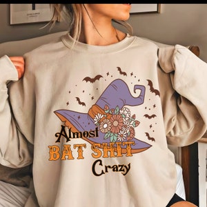 May include: Beige sweatshirt with a graphic of a purple witch hat adorned with flowers and bats. The text "Almost BAT SHIT Crazy" is printed below the hat in orange and black.
